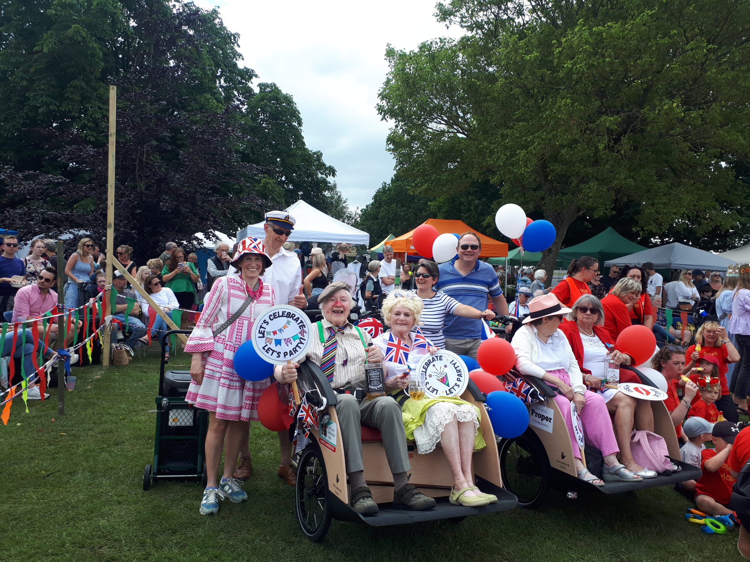 8 volunteers and passengers around 2 trishaws. They are at a fair, everyone is in fancy dress and there are lots of people and bunting in the background.