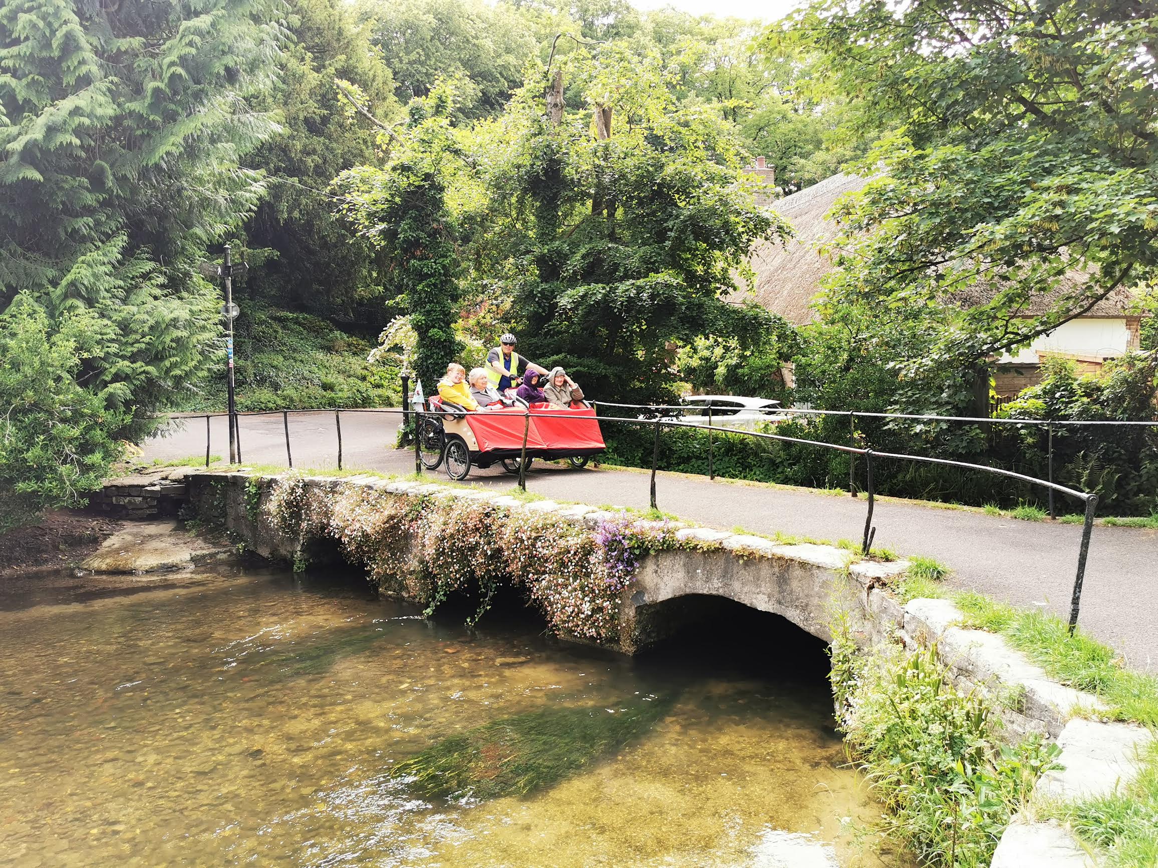 A trishaw with passengers and a volunteer pilot ride along a beautiful scenic bridge going over a river.