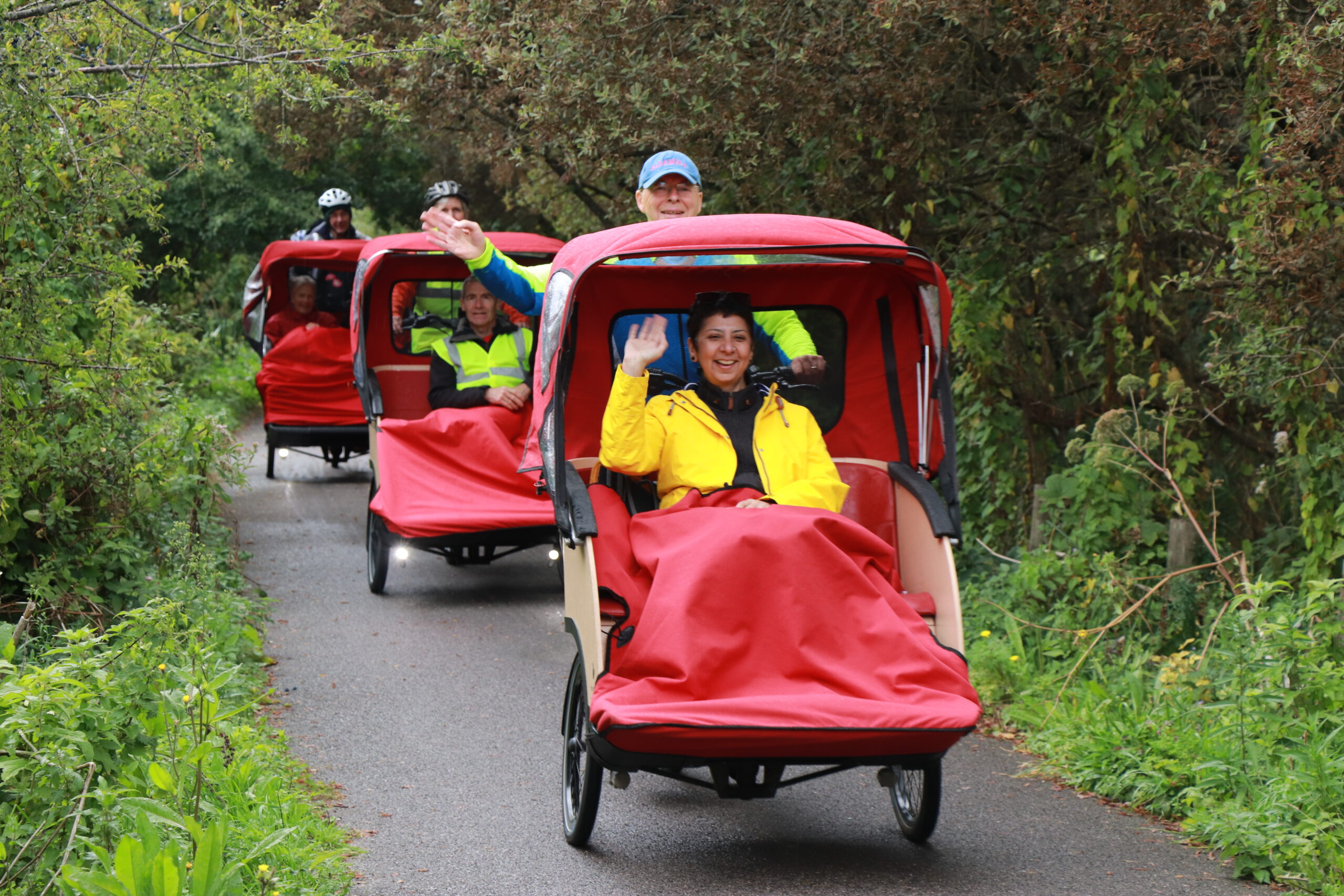 A woman in a bright yellow jacket sits in a red trishaw waving. She is on the move riding through a forest, you can see 3 more trishaws riding behind her.