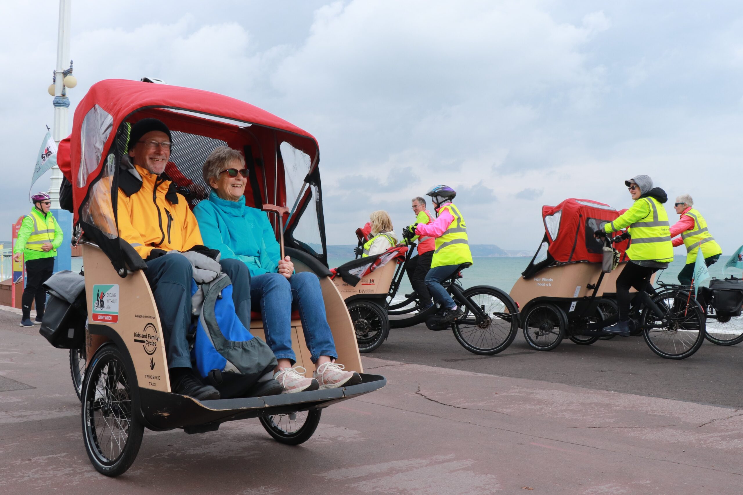 2 people sit in a trishaw smiling, behind them is 4 other red trishaws, volunteers in high vis jackets surround them