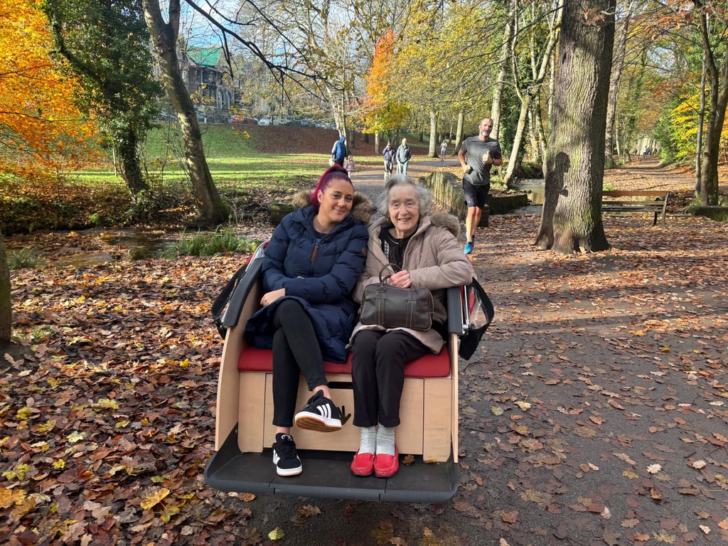 In a park in autumn a trishaw with an older and younger passenger sits on a brown leafy path.