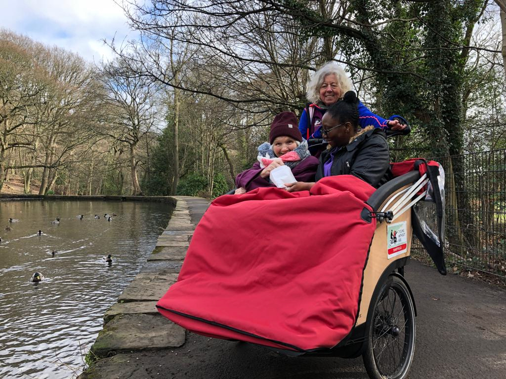 A trishaw with 2 passengers are wrapped up warmly in front of a pond in a park