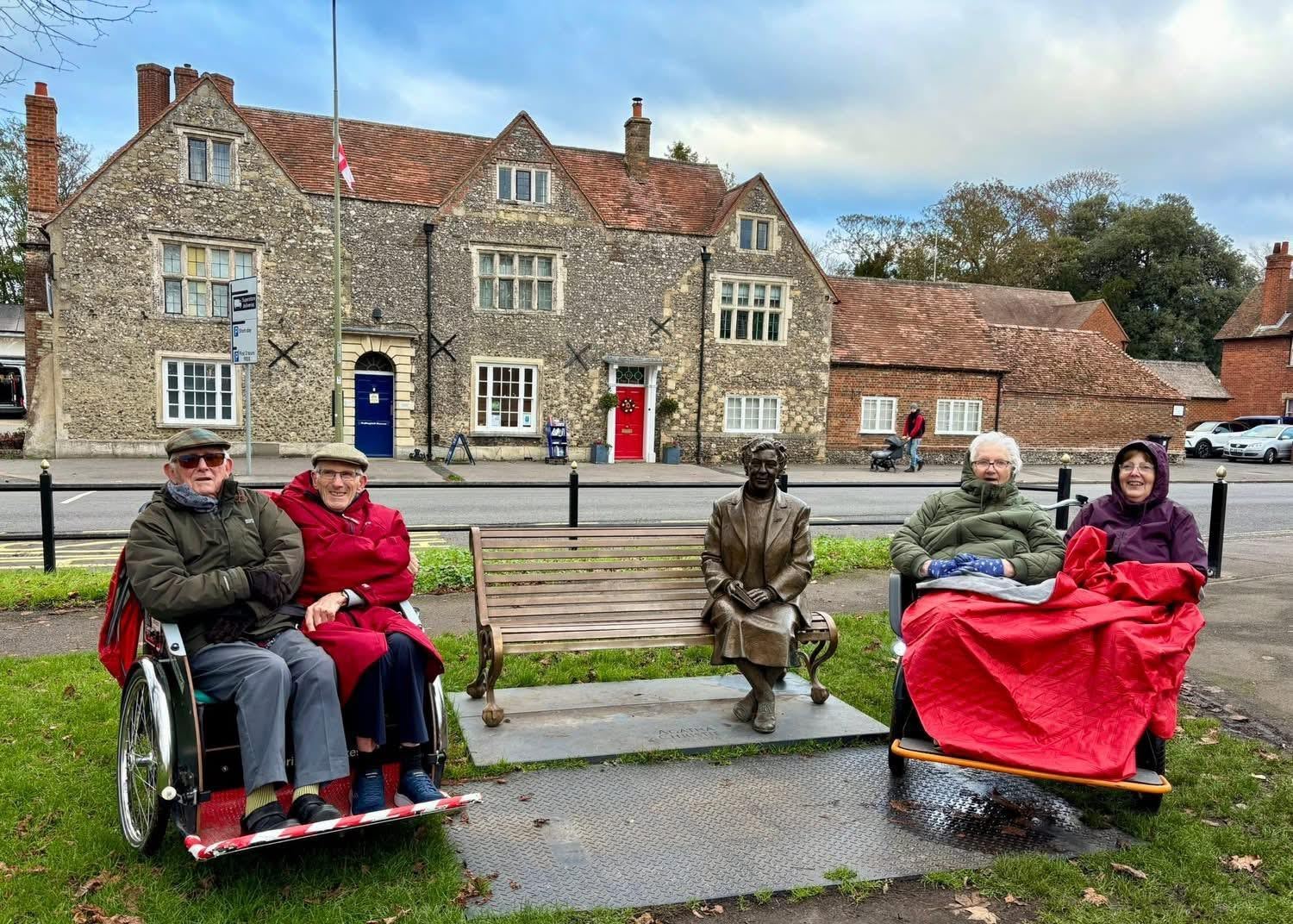 There are two trikes each with two passengers on them with a red blanket and gloves. They are sat on either side of a bench with a statue of Agatha Christie