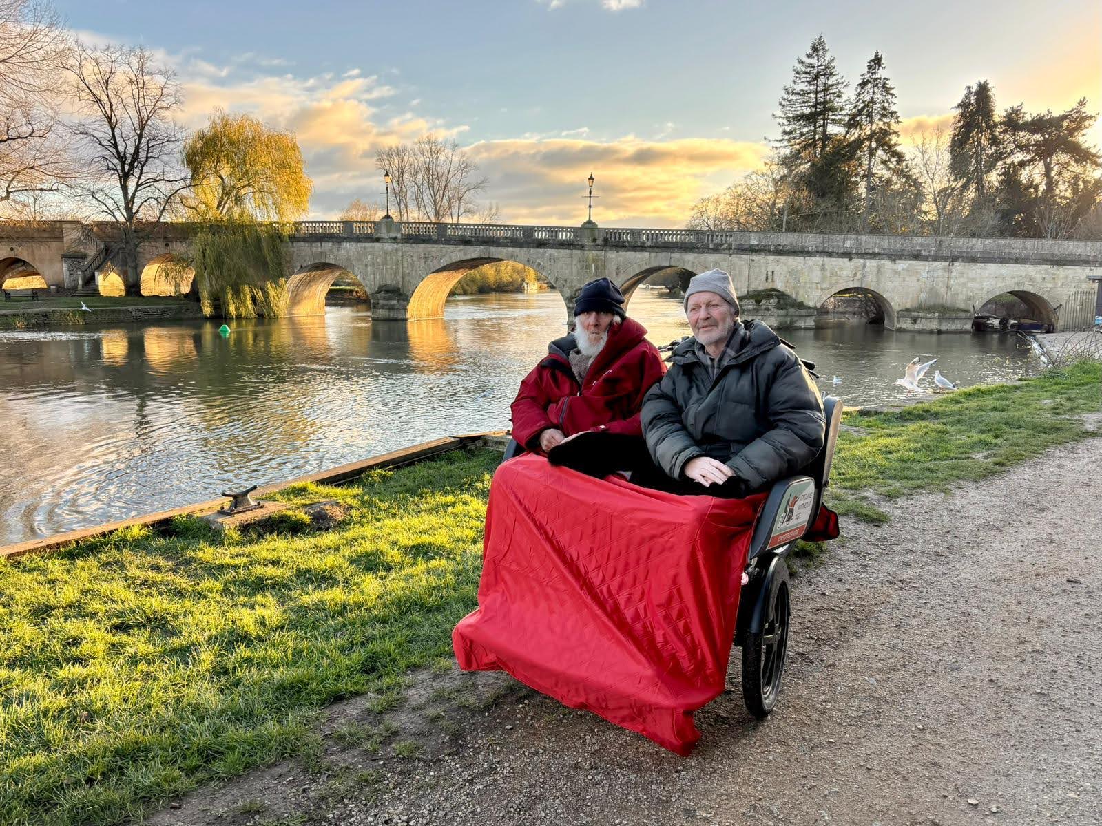 Two male passengers are sat on a trike with a red blanket with the river Thames behind them and Wallingford bridge.